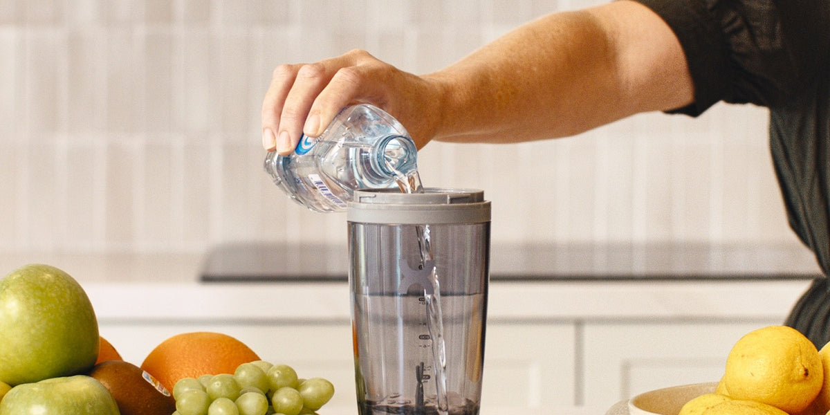 A hand in a black sleeve pours water into an electric shaker on a marble bench, with berries, apples, lemons and eggs in bowls — HLTH+
