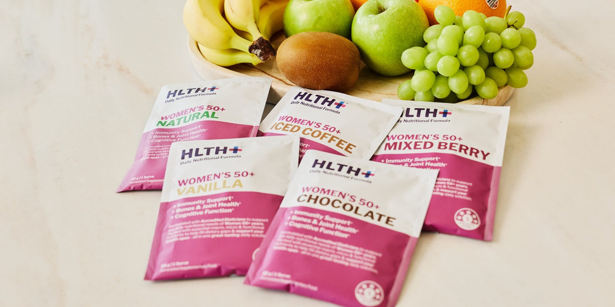 Five HLTH+ Women's 50+ sachets in pink arranged on a light marble benchtop with fresh fruit behind — HLTH+