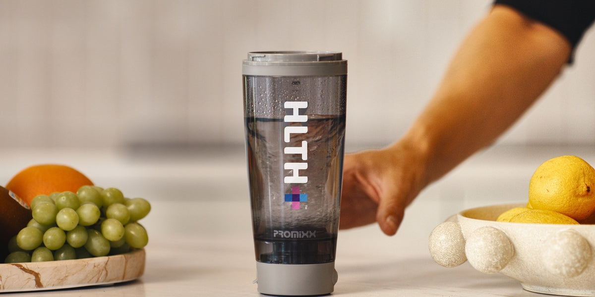 A HLTH+ electric shaker on a light stone benchtop with green grapes in a cork dish, lemons and an orange — HLTH+