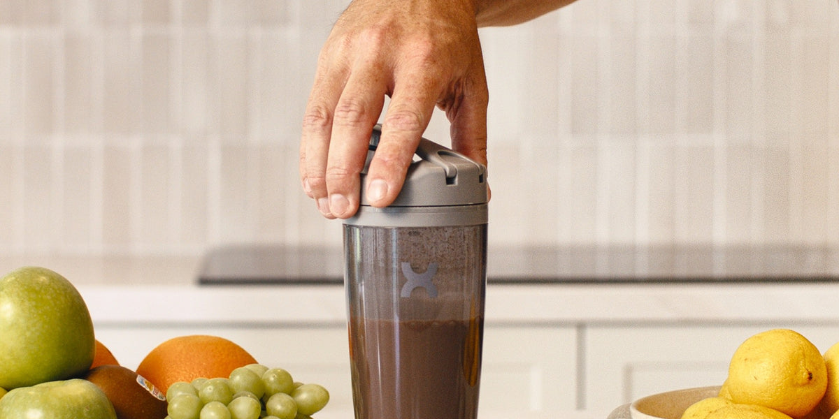 A man's hand pressing the button on a charcoal electric shaker on a white marble bench, fruit in wooden bowls either side — HLTH+