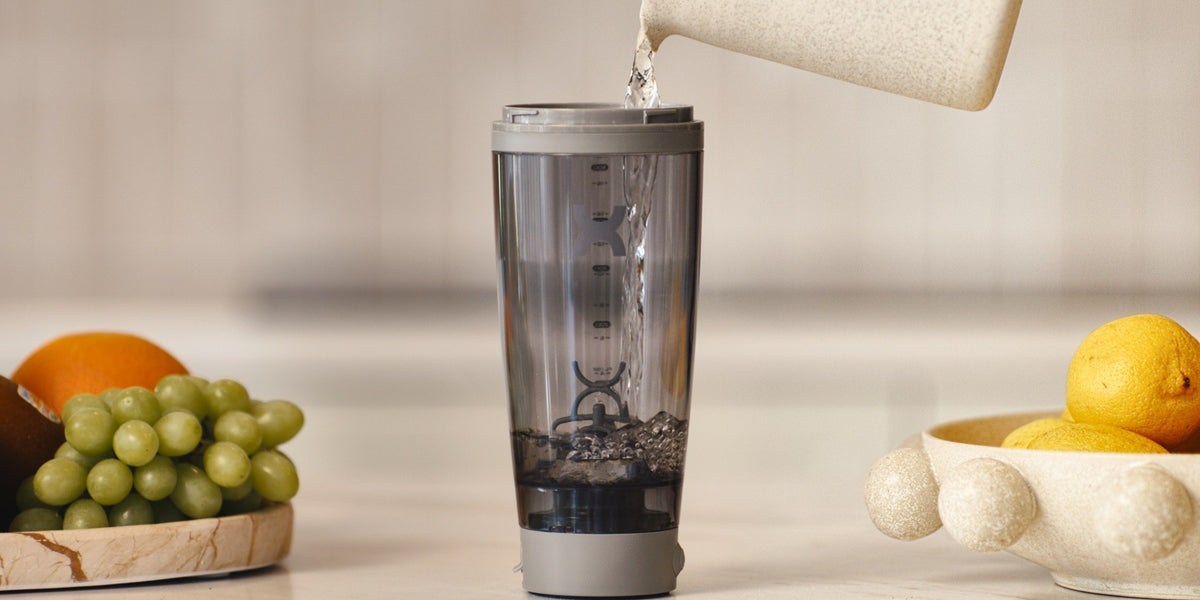 A HLTH+ electric shaker mixing powder into water, mid-pour from a sachet held above — HLTH+
