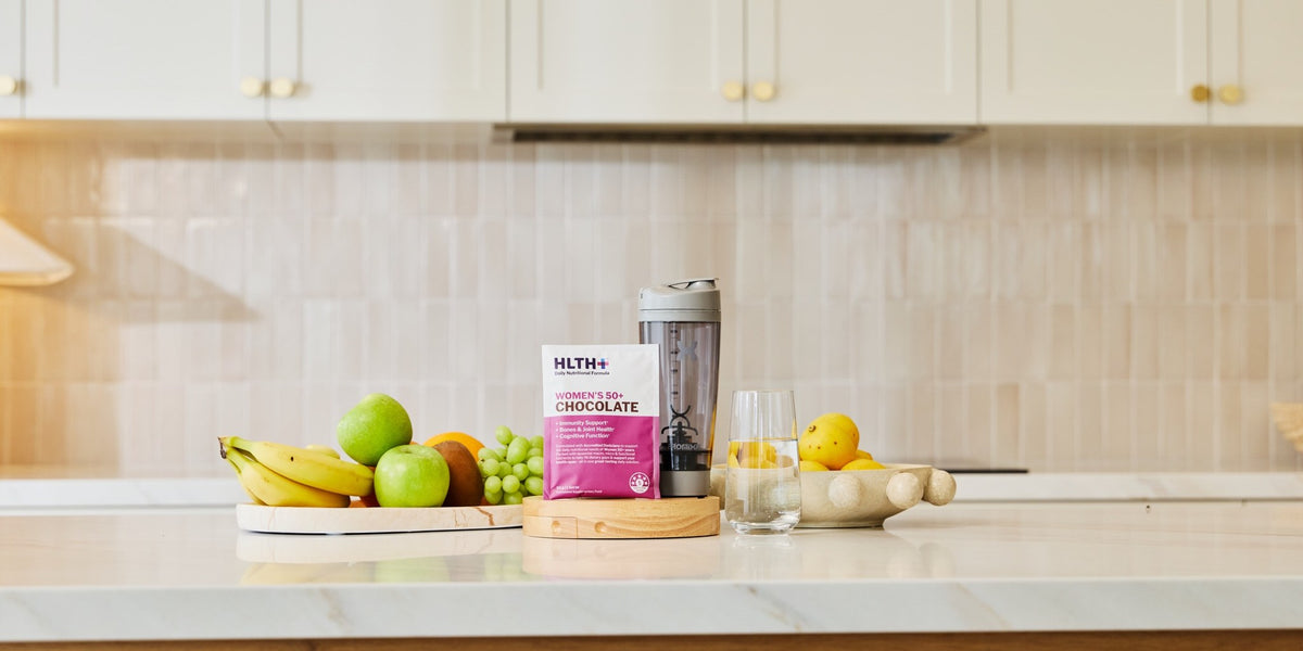 A white marble kitchen benchtop with a HLTH+ Women's 50+ sachet, cream cabinetry and vertical tile behind — HLTH+