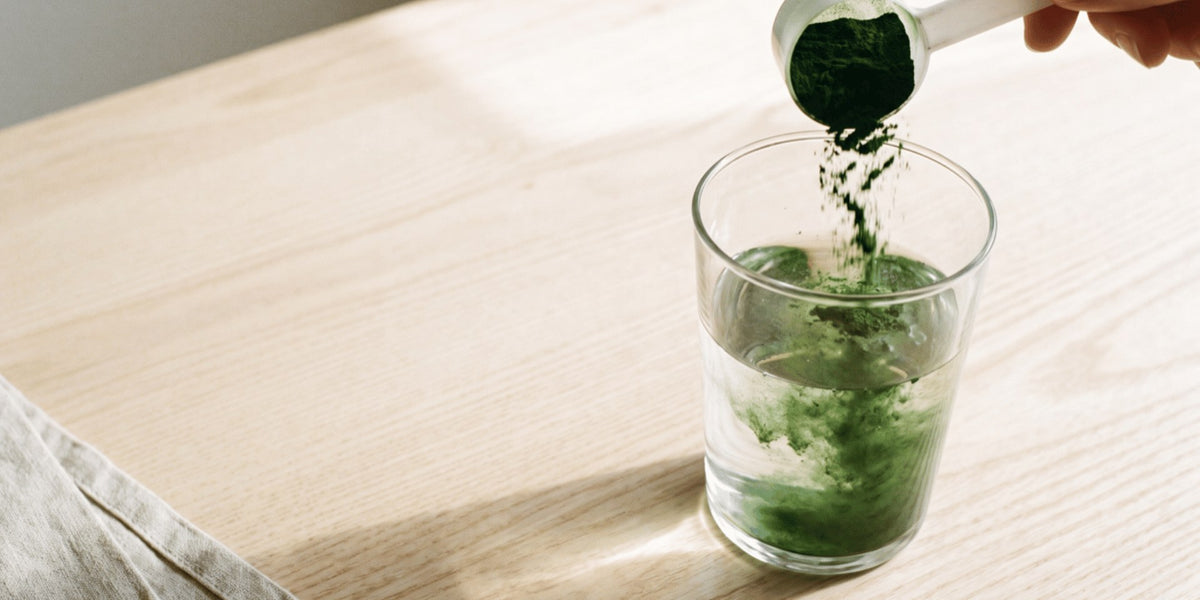Green powder poured from a scoop into a glass of water, shot top-down on a pale wooden bench — HLTH+