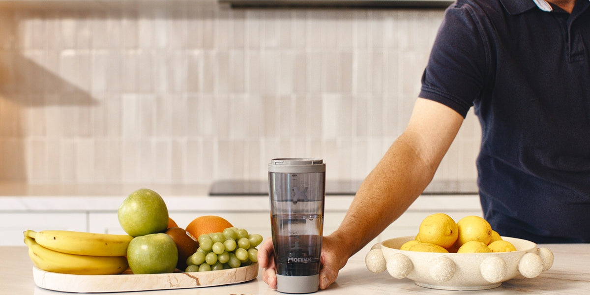 A man in a navy polo holding an electric shaker in a modern kitchen with a white brick backsplash — HLTH+