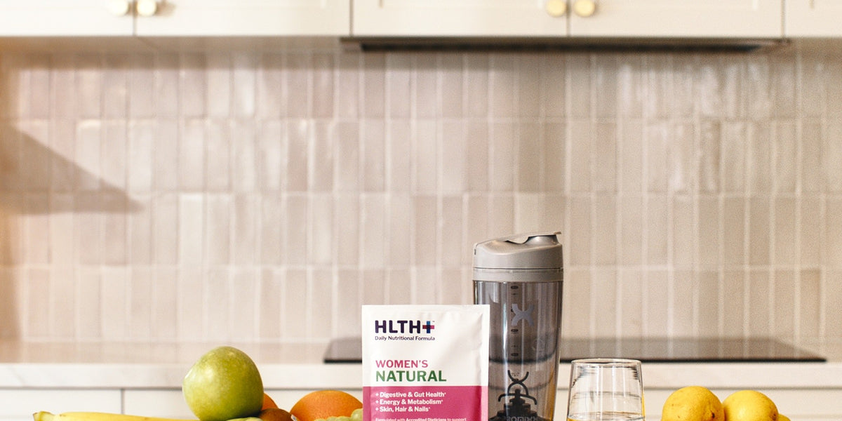 HLTH+ Women's Natural sachet with a charcoal electric shaker, a glass of water and bowls of fresh fruit on a kitchen bench — HLTH+