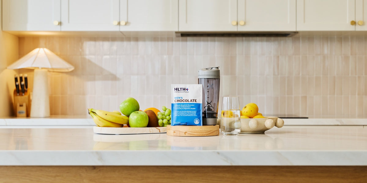 A HLTH+ Men's Chocolate sachet on a marble benchtop in a modern kitchen with an electric shaker — HLTH+