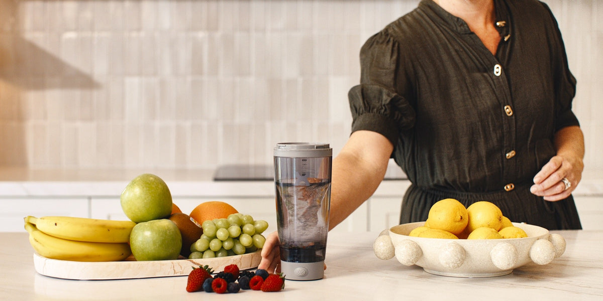A woman in her 50s stands behind a bright kitchen bench with bananas, berries and lemons, a black electric shaker and a glass beside her — HLTH+