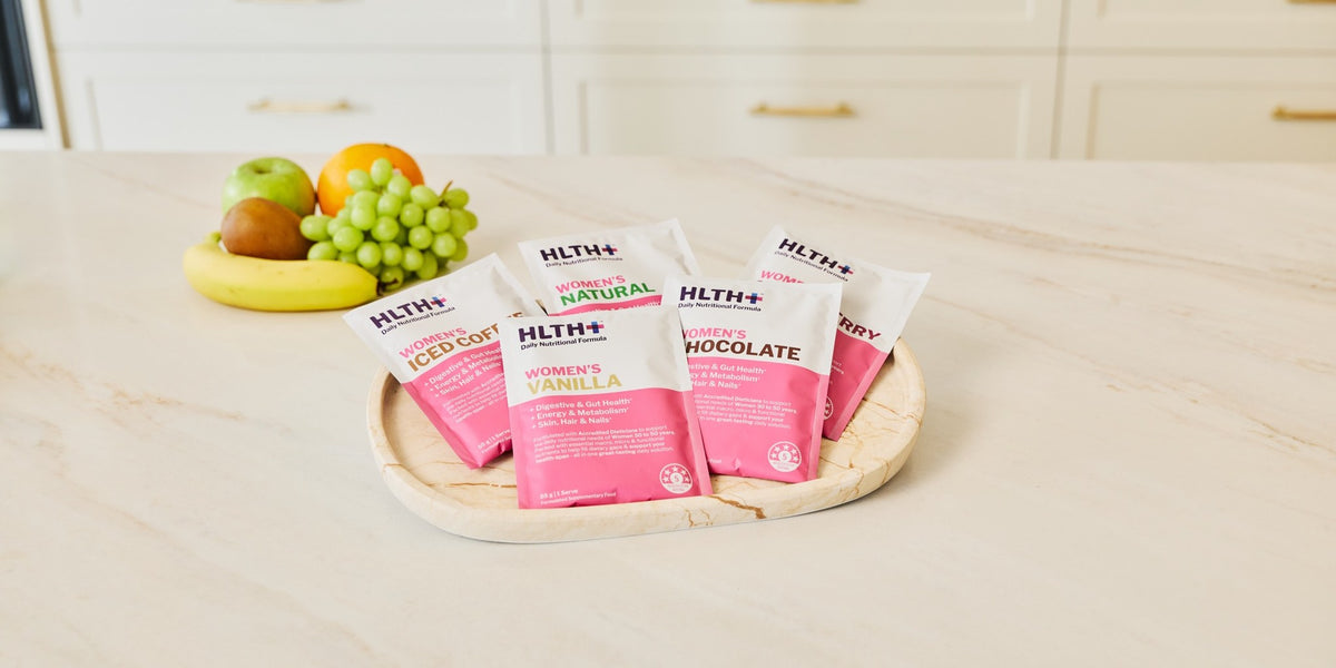 Five pink HLTH+ Women's sachets in Natural, Vanilla and Chocolate arranged on a round wooden coaster on a marble bench, with fruit behind — HLTH+