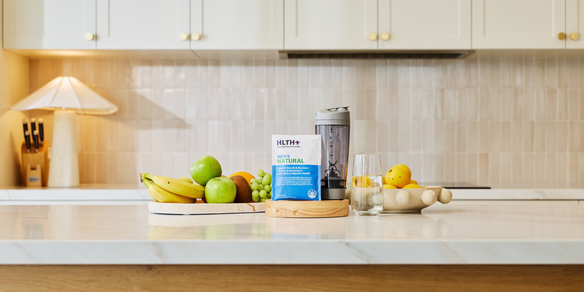 A HLTH+ Men's Natural sachet on a marble kitchen bench with fresh produce, a blender and a glass of water under a warm pendant lamp — HLTH+