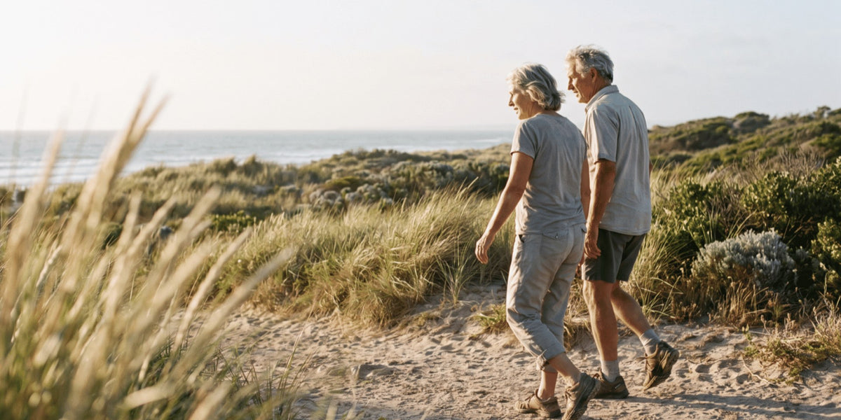 Two people in their sixties walking a sandy coastal path in the late afternoon — HLTH+