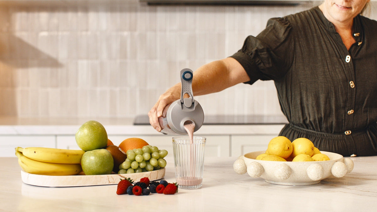 Woman pouring a HLTH+ shake from an electric shaker into a glass beside a tray of fresh fruit