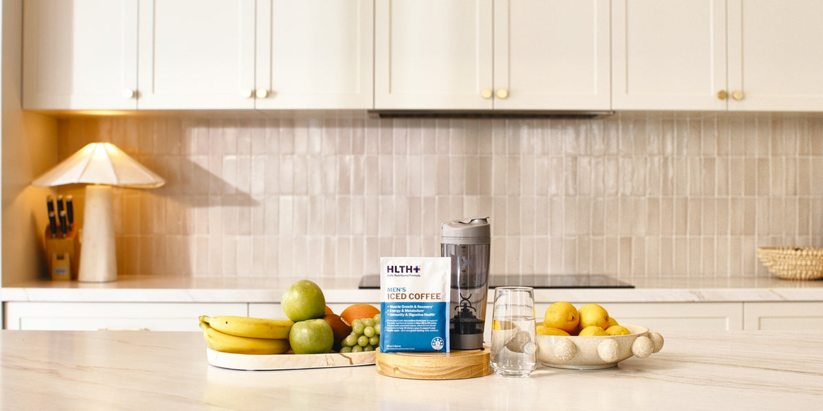 A HLTH+ Women's Iced Coffee sachet on a light wood kitchen benchtop beside an electric shaker — HLTH+