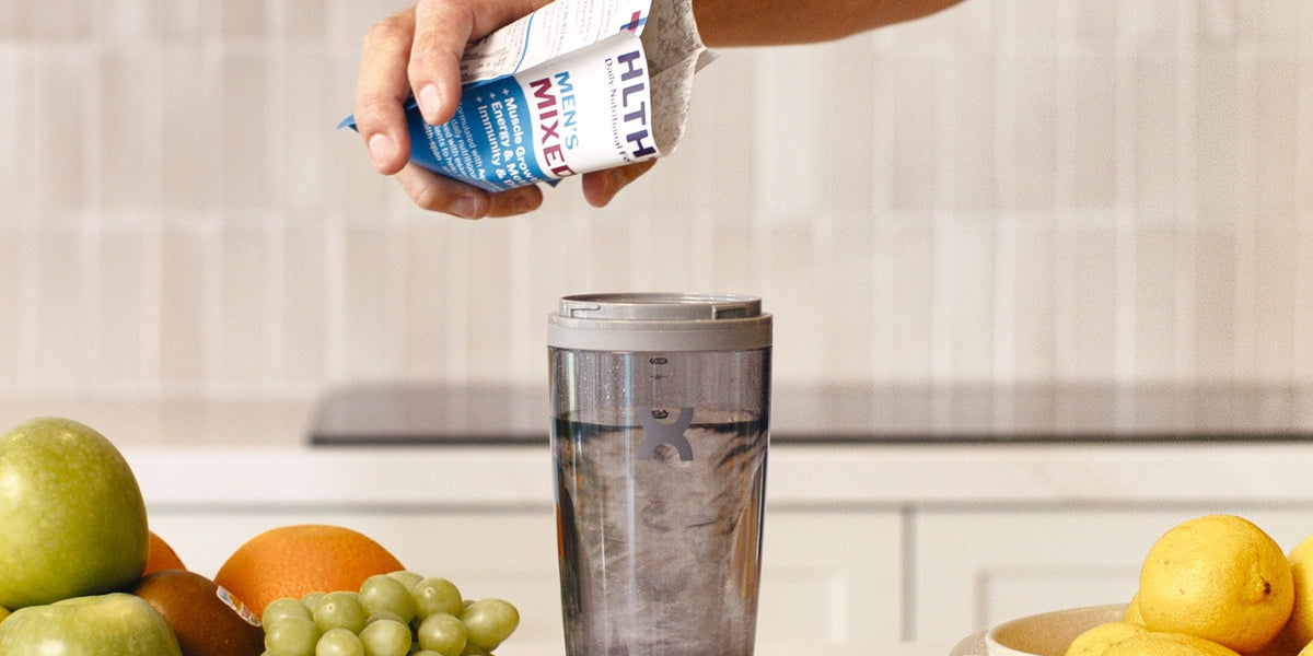 A hand pours a blue HLTH+ Men's sachet into an electric shaker of water above a kitchen bench scattered with fresh fruit and berries — HLTH+