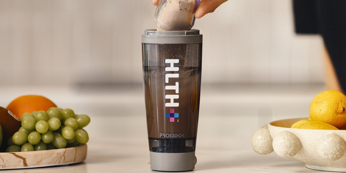 A hand pouring a sachet into a HLTH+ electric shaker on a white bench with fresh fruit beside it — HLTH+