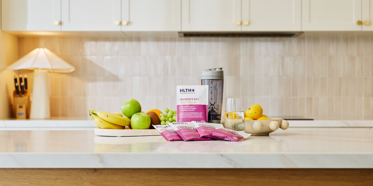 A white marble kitchen benchtop with HLTH+ Women's 50+ sachets in pink packaging and cream cabinetry behind — HLTH+