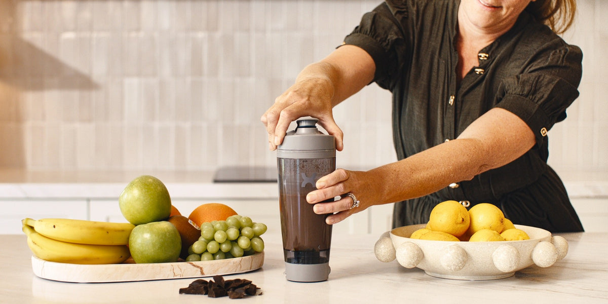 A woman in her 50s smiling as she uses a charcoal electric shaker at a light wood kitchen bench with fresh fruit in front of her — HLTH+