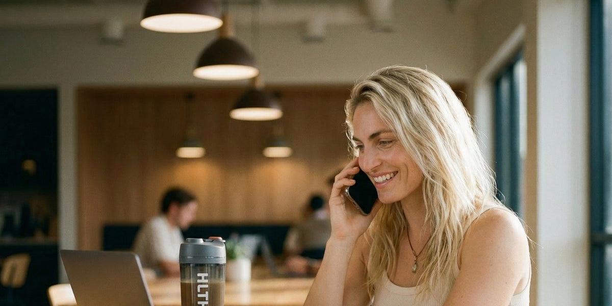 A woman at a wooden office desk on a phone call, a HLTH+ sachet and shaker on the desk beside her — HLTH+