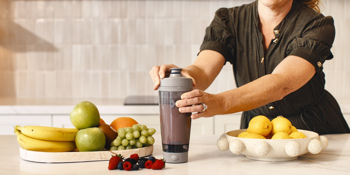A woman in her fifties holding a HLTH+ electric shaker over a kitchen bench laid with bananas, apples and grapes — HLTH+