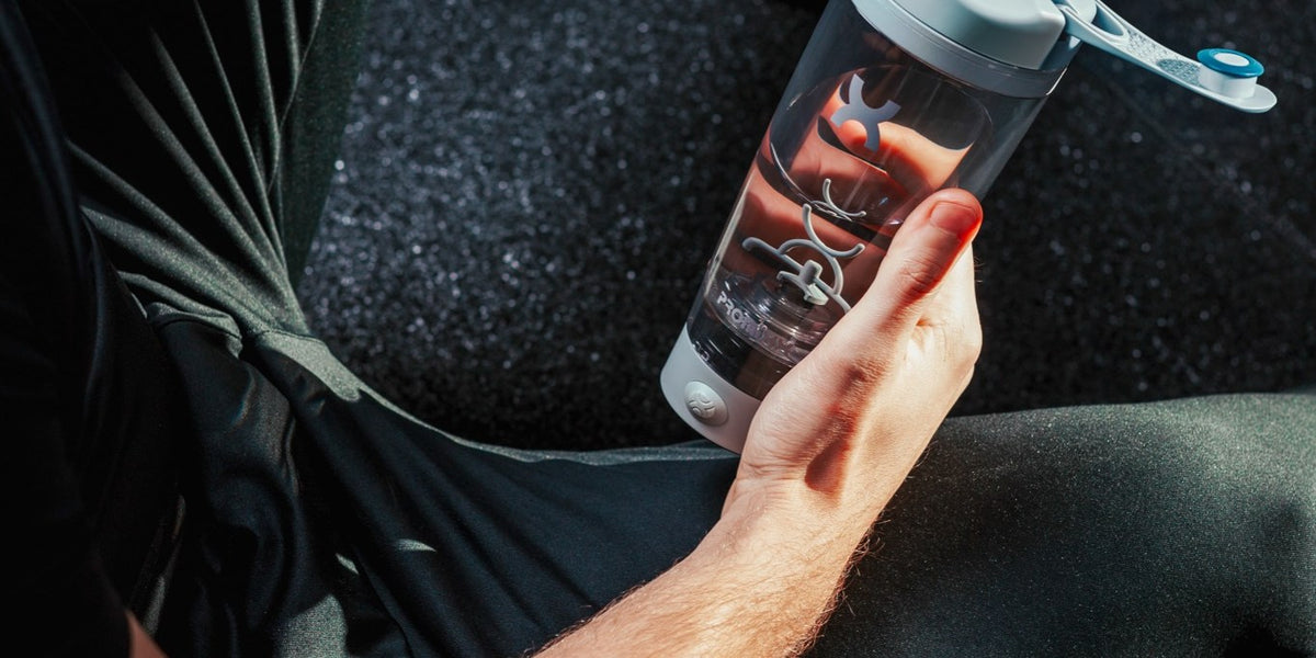 A person in black activewear sits on a gym floor in warm sunlight, mixing a drink in an electric shaker — HLTH+