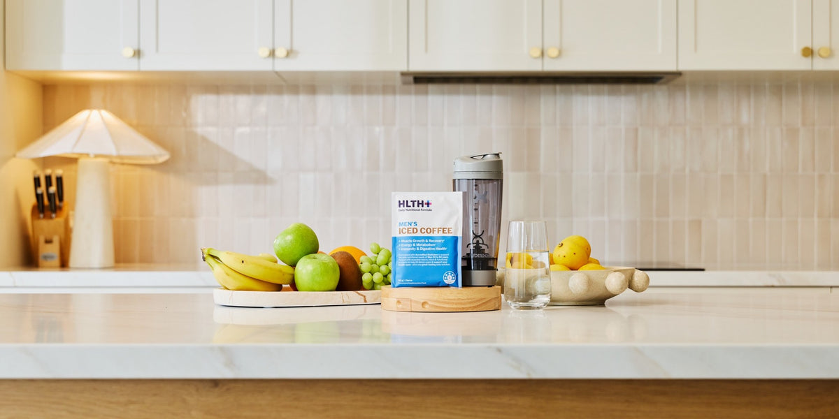 HLTH+ Men's Iced Coffee sachet beside a black electric shaker on a white marble kitchen bench with bananas, apples and grapes — HLTH+