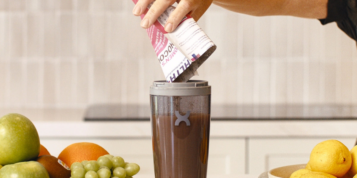 A woman's hand pouring a pink HLTH+ sachet into a charcoal electric shaker on a white bench with fresh fruit around it — HLTH+