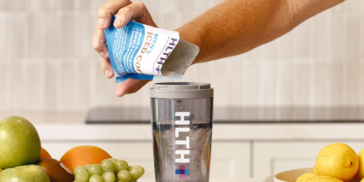 A man's hand pouring an HLTH+ Men's Daily Nutritional Formula sachet into a charcoal HLTH+ Pro Electric Shaker on a white bench — HLTH+