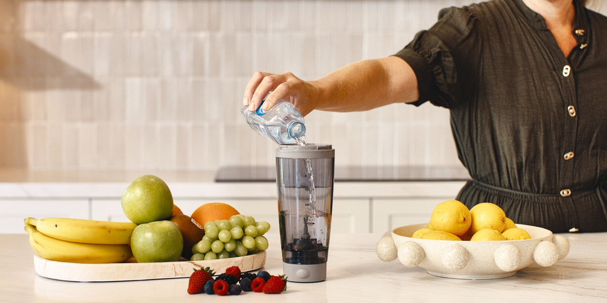 A woman pouring water into a HLTH+ electric shaker on a wooden kitchen bench with bananas, apples and berries beside it — HLTH+