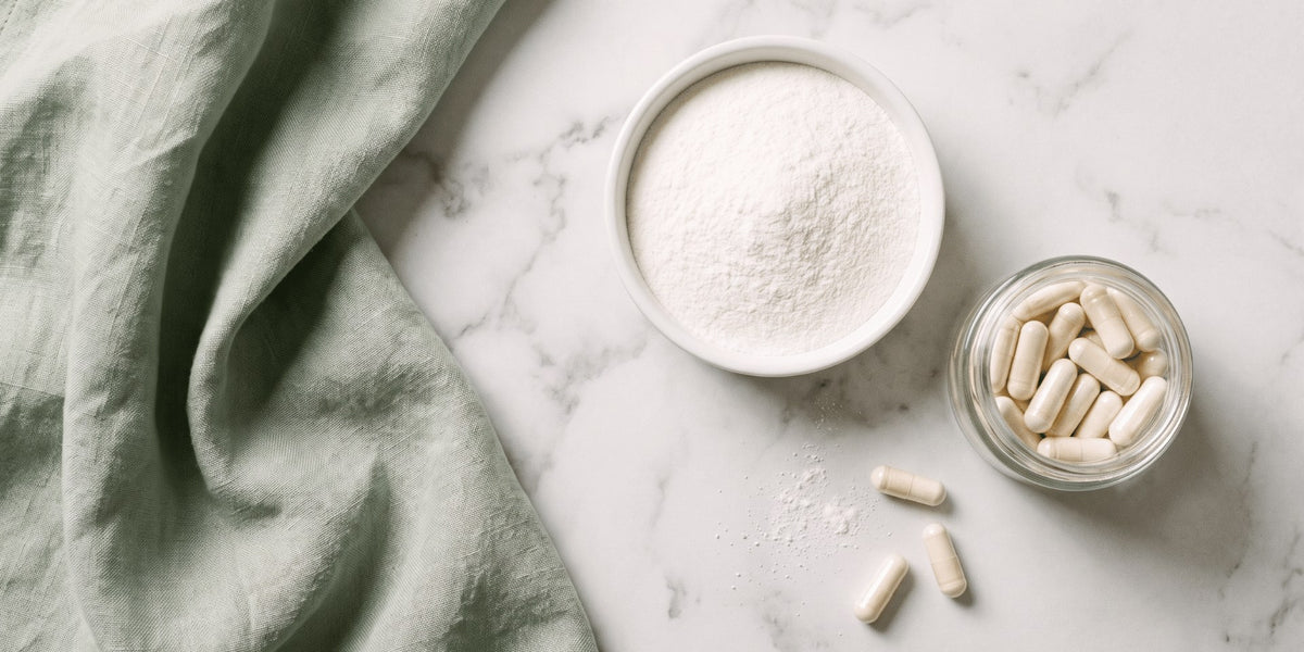 A small bowl of white magnesium powder beside a jar of capsules on a marble bench with green linen — HLTH+