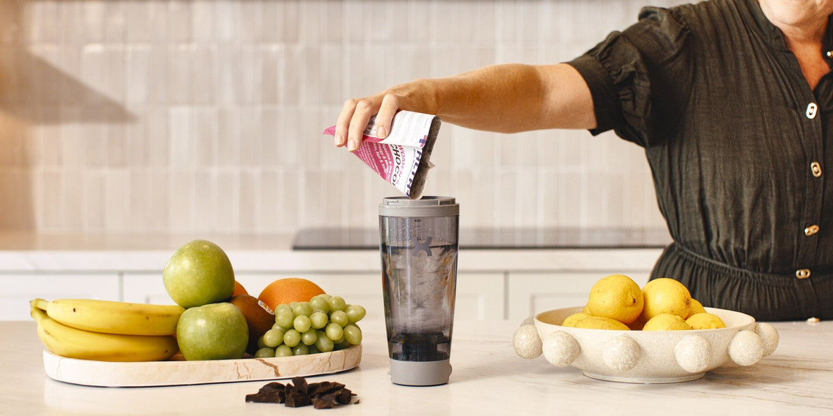 A woman in her 50s pours a pink HLTH+ Women's 50+ sachet into an electric shaker of water on a light wood kitchen bench, with bananas, apples and grapes beside her — HLTH+