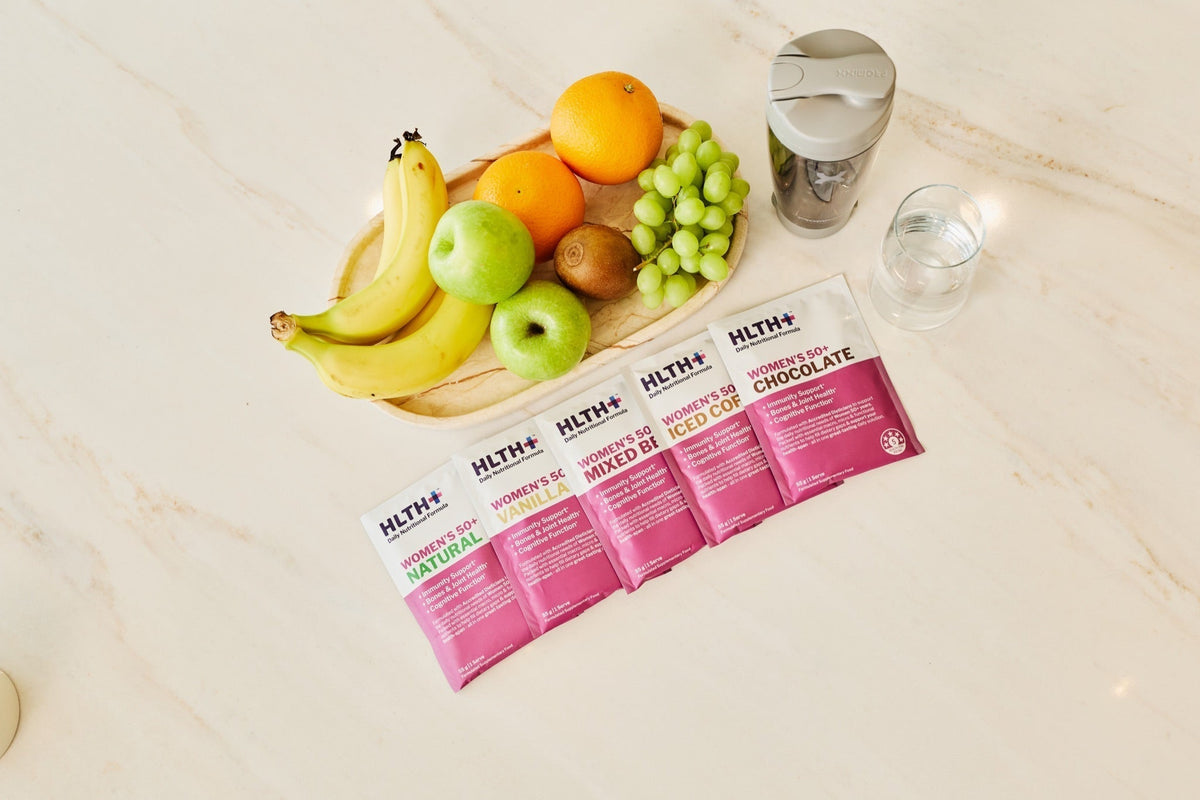 Five HLTH+ Women's 50+ sachets in all five flavours laid out with an electric shaker, water and fresh fruit