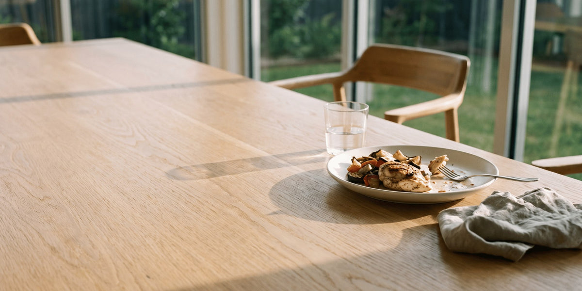 A half-eaten plate of food pushed to one side of a wooden dining table in natural light — HLTH+